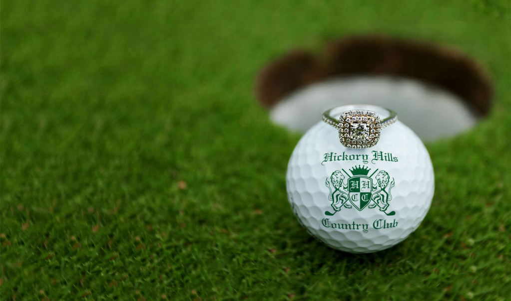 Branded golf ball with wedding ring balanced on top near golf course hole